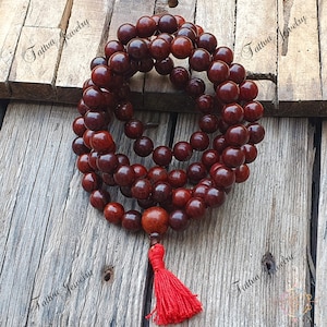May include: A red wooden mala bead necklace with a red tassel. The beads are round and have a dark red color. The necklace is on a wooden surface.