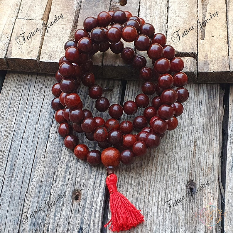 May include: A red wooden mala bead necklace with a red tassel. The beads are round and have a dark red color. The necklace is on a wooden surface.