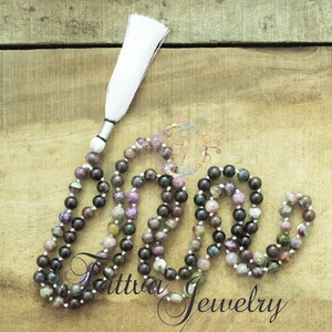 May include: A multi-colored gemstone mala necklace with a white tassel. The necklace is made of round beads with a mix of purple, pink, green, and black colors. The tassel is attached to the end of the necklace with a silver bead.