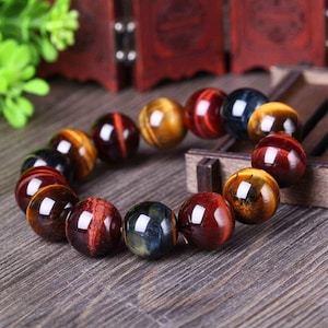 May include: A beaded bracelet featuring large, polished tiger eye stones in shades of brown, gold, red, and blue. The bracelet is strung together, showcasing the natural patterns and luster of the gemstones. The bracelet is displayed on a wooden surface.