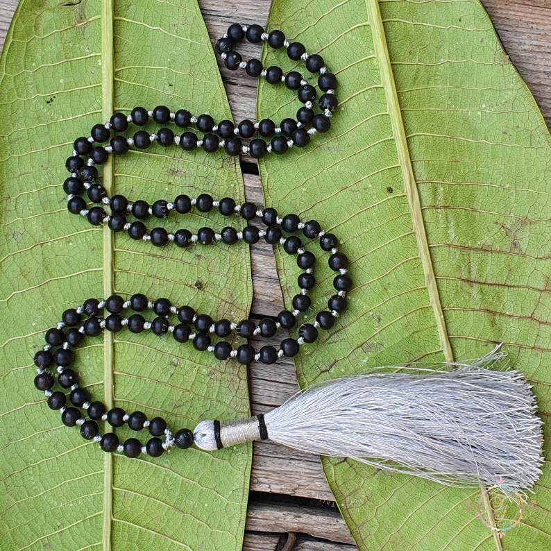 May include: A black beaded necklace with a silver tassel. The necklace is made of round beads and is strung on a silver cord. The tassel is made of silver thread and is attached to the end of the necklace.