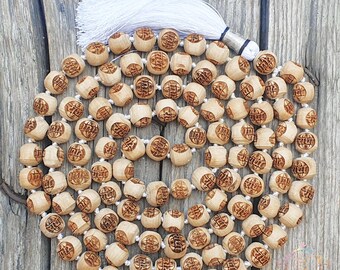 May include: A wooden mala bead necklace with a white tassel. The beads are round and have a carved design. The necklace is laid out on a wooden surface.