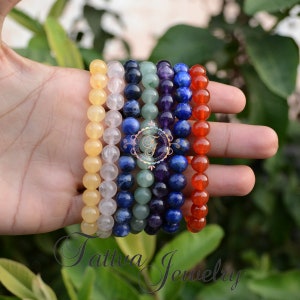 Può includere: Sette braccialetti dei chakra impilati su una mano. I braccialetti sono fatti di varie pietre preziose di diversi colori, tra cui giallo, bianco, blu, verde, viola e rosso.