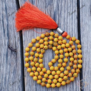 Puede incluir: Un collar de cuentas mala amarillo con una borla naranja. Las cuentas están ensartadas en un hilo naranja. La borla es de color naranja brillante con un acento plateado y negro. El collar se exhibe sobre una superficie de madera envejecida.