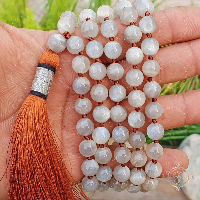 May include: A white moonstone mala with a tassel made of orange thread. The mala is made of round beads and is strung on a brown cord.