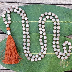 May include: A white moonstone mala with an orange tassel. The mala is made of 108 beads and is strung on a red cord.