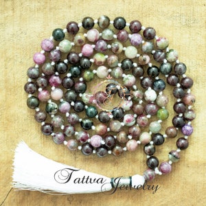 May include: A mala or prayer beads made of green, pink, and black gemstone beads with a white tassel. The beads are strung on a white cord. The mala is on a wooden surface. "Tattva Jewelry" is written on the surface.