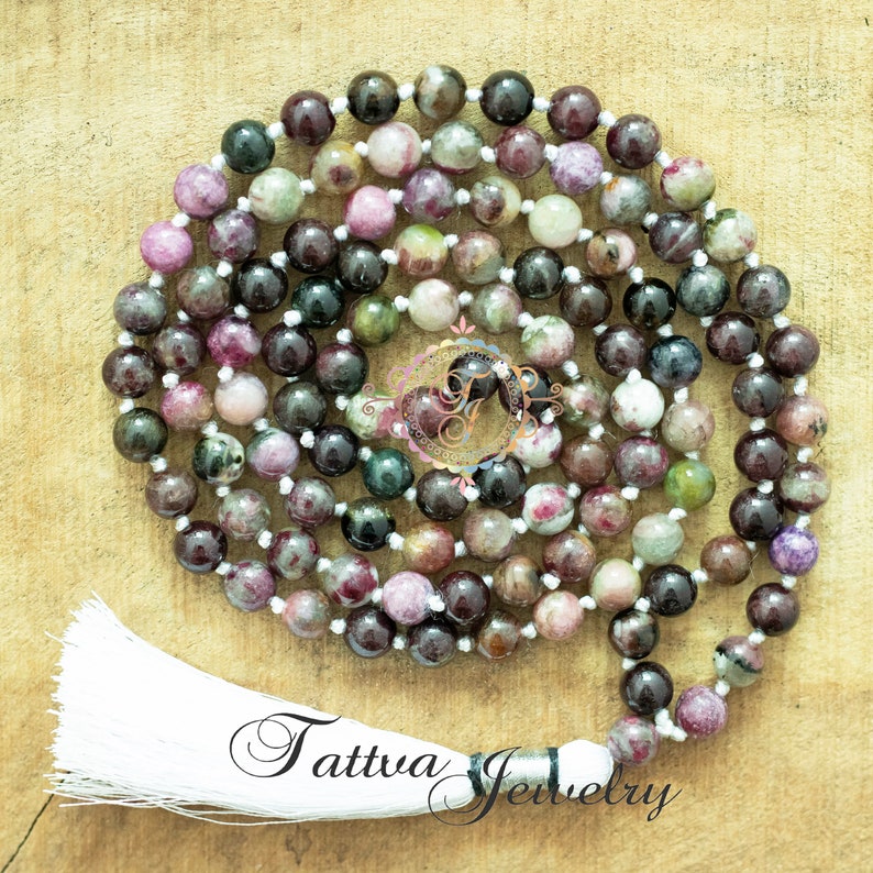 May include: A mala or prayer beads made of green, pink, and black gemstone beads with a white tassel. The beads are strung on a white cord. The mala is on a wooden surface. "Tattva Jewelry" is written on the surface.