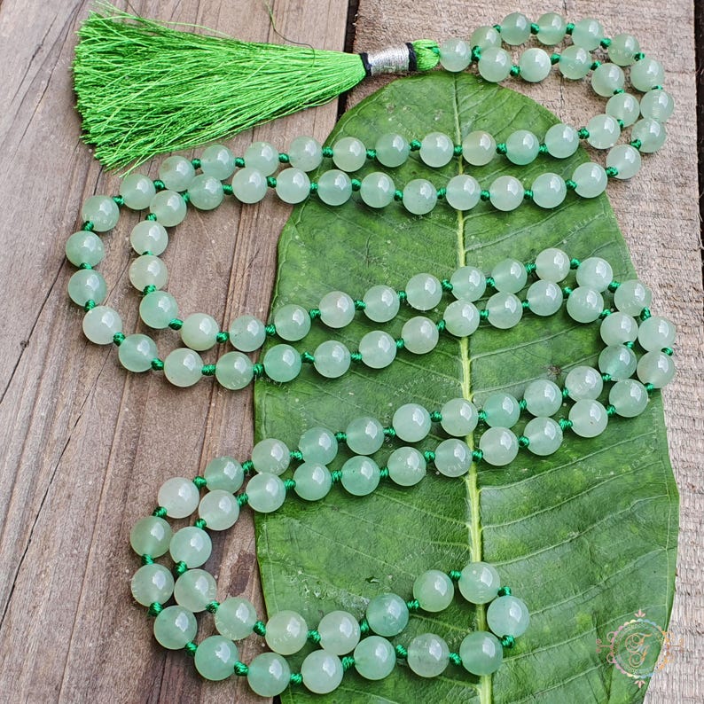 May include: A green jade mala with a green tassel. The mala is made of round beads and is strung on a green cord. The mala is lying on a green leaf.