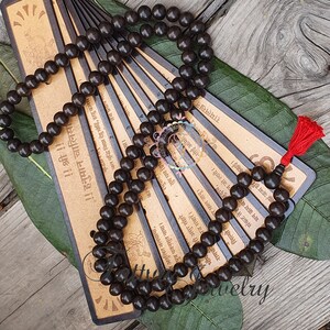 May include: A dark brown wooden mala bead necklace with a red tassel. The beads are strung on a cord and the necklace is laid out on a wooden surface with a set of cards with text in a foreign language.