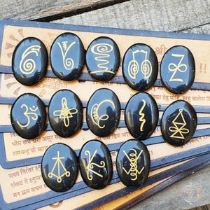 May include: Twelve black, oval stones with gold symbols, arranged on a light brown surface. The symbols are diverse, featuring spirals, lines, and geometric shapes. The background shows some text. The stones are likely used for spiritual or decorative purposes.