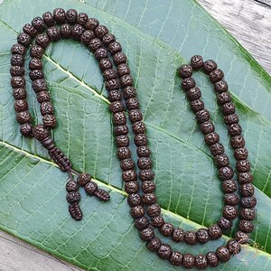 May include: A brown wooden mala bead necklace with 108 beads. The beads are round and have a textured surface. The necklace is lying on a green leaf.