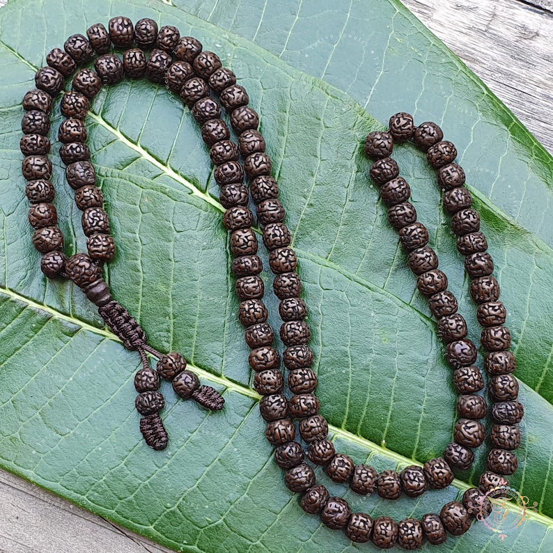 May include: A brown wooden mala bead necklace with 108 beads. The beads are round and have a textured surface. The necklace is lying on a green leaf.