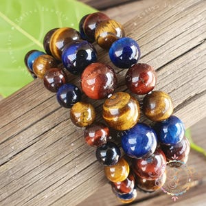 May include: A multi-strand bracelet composed of polished, spherical gemstone beads in shades of deep blue, golden brown, and reddish-brown. The beads are arranged in a repeating pattern, showcasing natural variations in colour and lustre throughout.