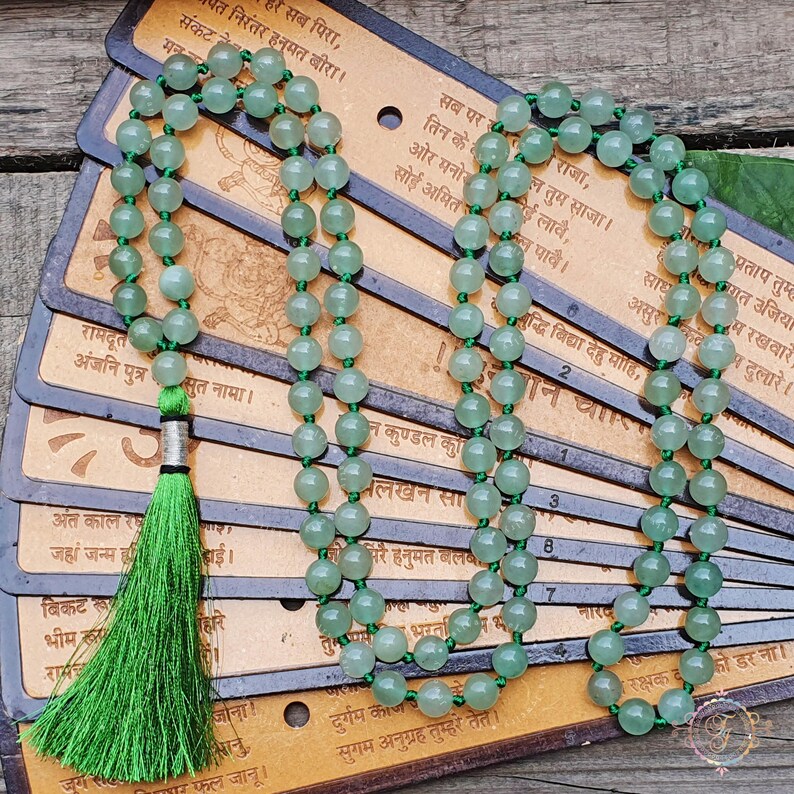 May include: A green mala necklace with 108 beads, a green tassel, and a silver charm. The necklace is lying on a stack of cards with Sanskrit text.