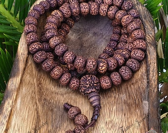 May include: A brown wooden mala bead necklace with 108 beads. The beads are round and have a textured surface. The necklace is on a wooden surface.