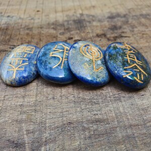 May include: Four smooth, blue and gold stones with different symbols etched on them. The stones are arranged in a row on a wooden surface.