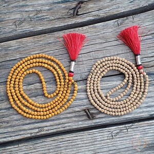 May include: Two malas, or prayer beads, with red tassels. One mala is made of light brown beads and the other is made of yellow beads. Both malas are laid out on a wooden surface.