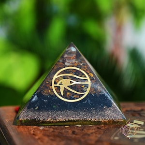 Könnte beinhalten: Eine schwarze Obsidianpyramide mit einem goldenen Horusauge-Symbol und einem klaren Quarzkristall an der Spitze. Die Pyramide ist mit kleinen braunen und goldenen Kristallen gefüllt.