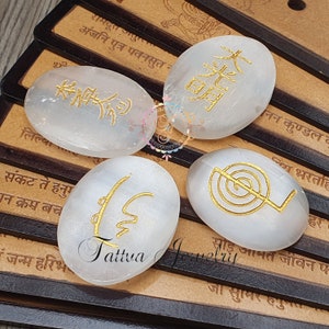 May include: Four white selenite stones with gold symbols and text. The stones are oval-shaped and arranged on a wooden surface with text. The text on the stones is in gold. The stones are approximately 5 cm long.