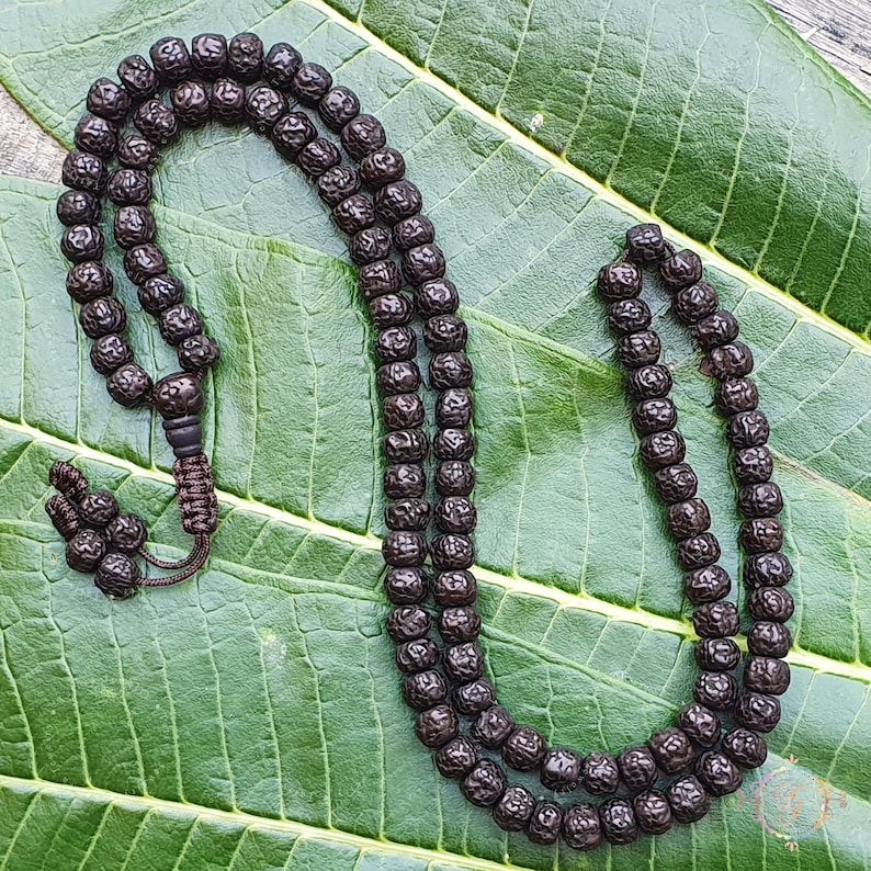 May include: A dark brown wooden mala bead necklace with 108 beads. The necklace is lying on a green leaf background.