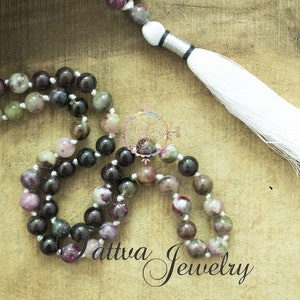 May include: A multi-colored gemstone mala necklace with a white tassel. The necklace is made of round beads with shades of pink, green, and black. The tassel is attached to a silver cord.