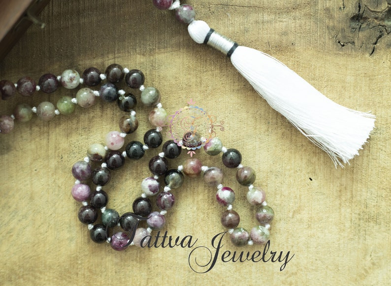 May include: A multi-colored gemstone mala necklace with a white tassel. The necklace is made of round beads with shades of pink, green, and black. The tassel is attached to a silver cord.