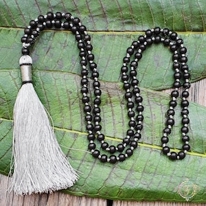 May include: A black beaded necklace with a silver tassel. The necklace is made of round beads and is strung on a black cord. The tassel is made of a soft, gray material.