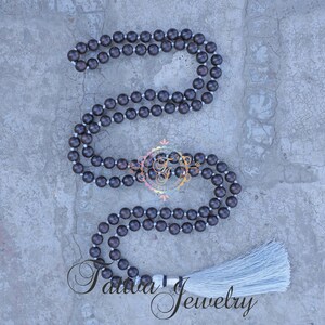 May include: A dark brown wooden mala necklace with a white tassel. The necklace is made of round beads and is approximately 108 beads long.