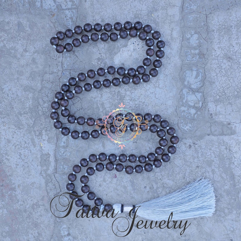 May include: A dark brown wooden mala necklace with a white tassel. The necklace is made of round beads and is approximately 108 beads long.