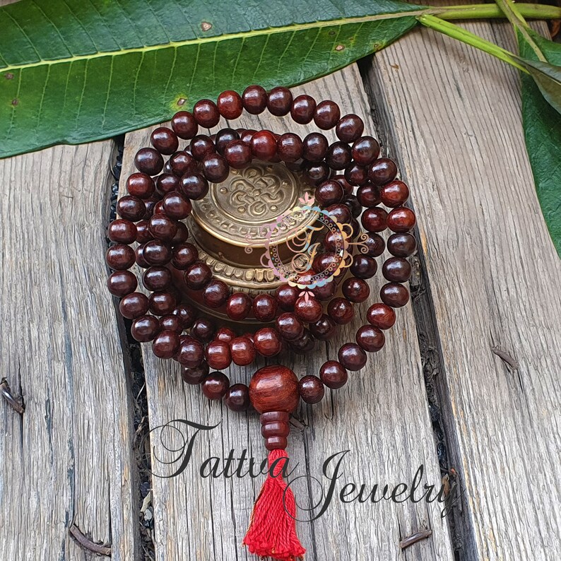May include: A dark brown wooden mala bead necklace with a red tassel. The mala beads are strung on a cord and have a decorative knot at the end. The mala is resting on a wooden surface.