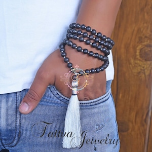 May include: A black beaded necklace with a silver charm and a white tassel. The necklace is wrapped around a person's wrist.