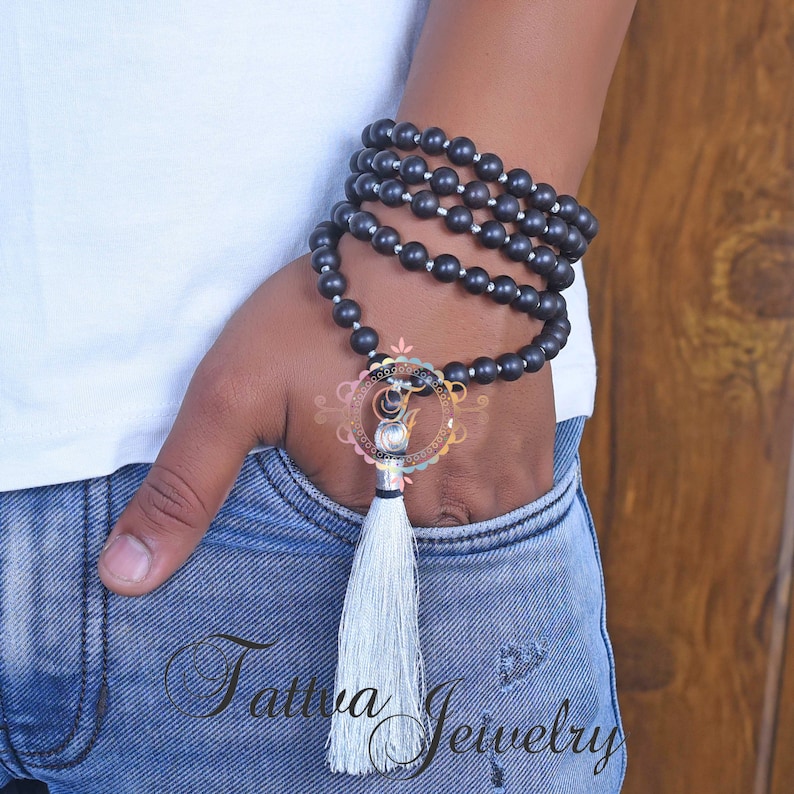 May include: A black beaded necklace with a silver charm and a white tassel. The necklace is wrapped around a person's wrist.