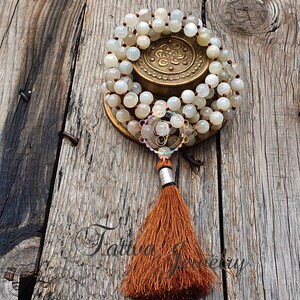 May include: A white moonstone mala with a brown tassel. The mala is on a wooden surface and is wrapped around a brass charm.
