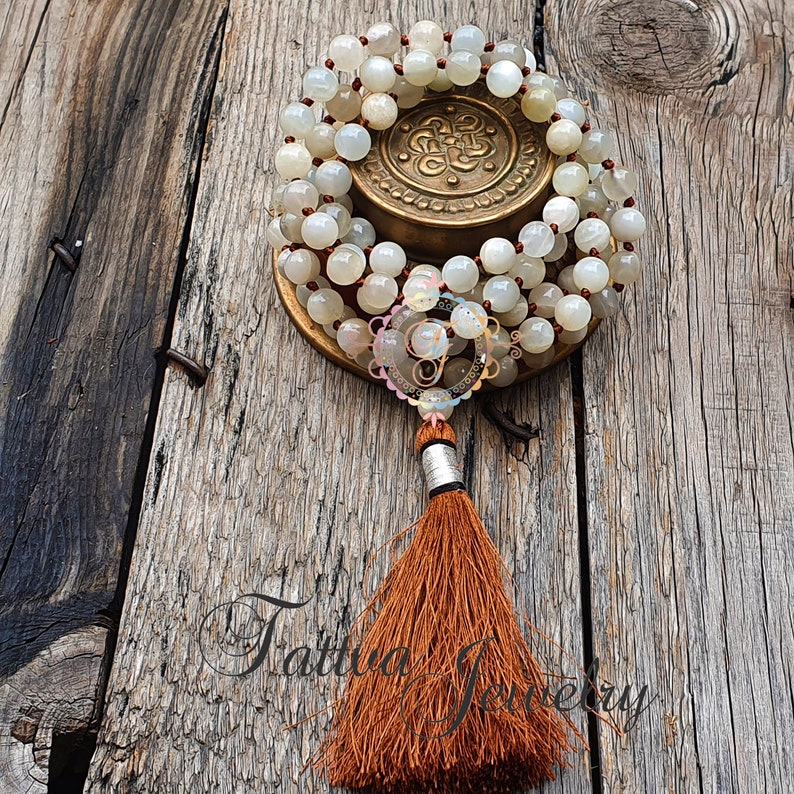 May include: A white moonstone mala with a brown tassel. The mala is on a wooden surface and is wrapped around a brass charm.