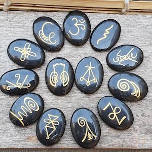 May include: Twelve black, oval stones with gold symbols, arranged on a wooden surface. The symbols are diverse, featuring abstract lines, spirals, and a musical note. The stones are a dark, glossy colour.