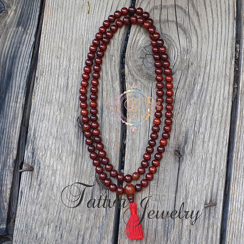May include: A dark red wooden mala bead necklace with a red tassel. The necklace is made of round beads and is approximately 108 beads long.