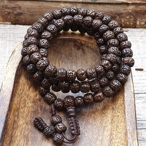 May include: A brown wooden mala bead necklace with 108 beads. The beads are round and have a textured surface. The necklace is tied with a brown cord.