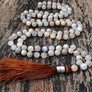 May include: A white moonstone mala necklace with a brown tassel. The necklace is made of round beads and is strung on a brown cord.
