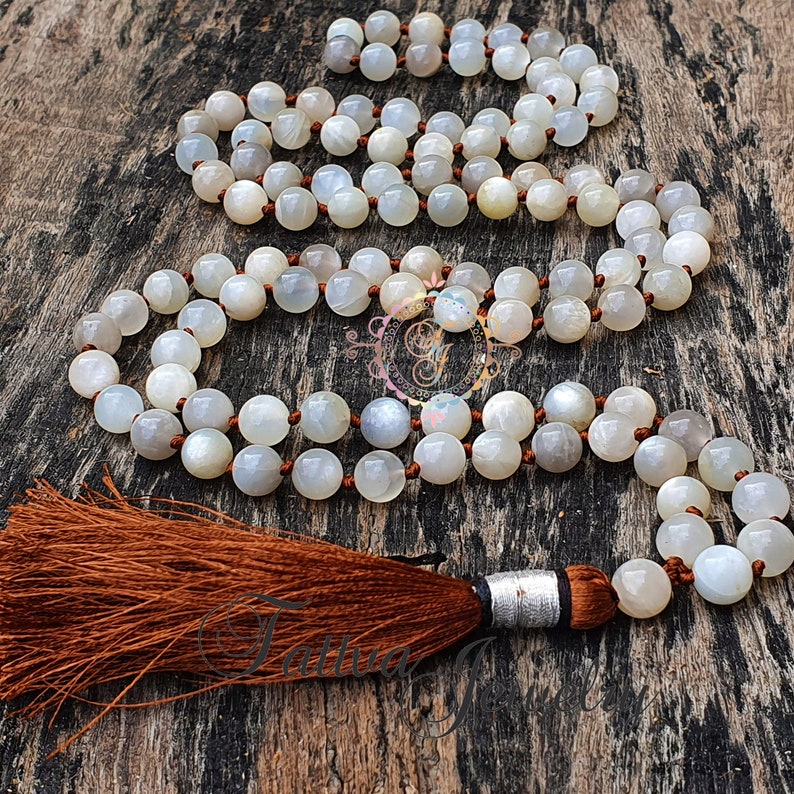 May include: A white moonstone mala necklace with a brown tassel. The necklace is made of round beads and is strung on a brown cord.
