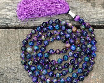 May include: A mala necklace with a purple tassel. The necklace is made of round beads in shades of blue, purple, and brown, strung on a purple cord. The tassel is made of purple threads and is attached to the necklace with a silver-coloured metal piece.