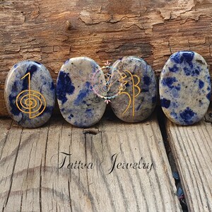 Puede incluir: Cuatro piedras lisas de forma ovalada, azules y blancas, con s&iacute;mbolos dorados grabados en la superficie. Las piedras est&aacute;n dispuestas en fila sobre una superficie de madera. El texto "Tattva Jewelry" es visible debajo de las piedras.