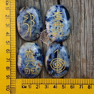 Puede incluir: Cuatro piedras lisas de forma ovalada de color azul con s&iacute;mbolos dorados. Las piedras miden aproximadamente 5 cm de largo y est&aacute;n dispuestas en una fila. Una regla es visible en el fondo.