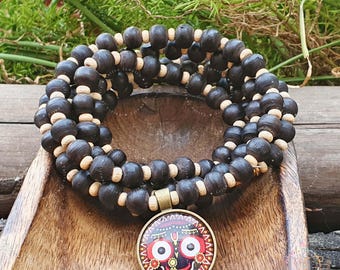 May include: A dark brown wooden bead wrap bracelet with a decorative pendant. The pendant features a colorful design with red, white, and black details. The bracelet is made of dark brown wooden beads and light brown spacers.