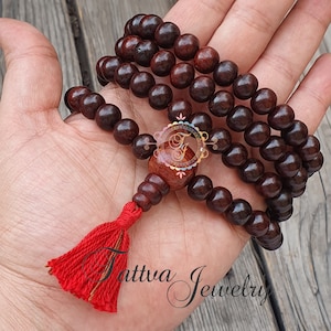 May include: A dark brown wooden mala with a red tassel. The mala has 108 beads and a larger bead at the end.