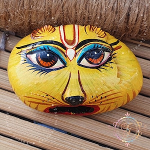 May include: A hand-painted, yellow stone sculpture of a cat's face. The cat's face has detailed features, including blue and orange eyes, black whiskers, and a red mouth. The sculpture is on a wooden surface.