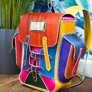 May include: A multi-coloured leather backpack with a yellow, orange, red, pink, blue, green, and purple colour scheme. The backpack has a large main compartment, a smaller front pocket, and two adjustable straps. The backpack has a metal buckle and a leather tag with the word "woff" on it.