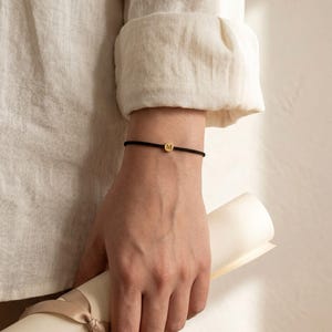 Puede incluir: Pulsera de cordón negro con un pequeño dije circular dorado con la letra "M". La pulsera se lleva en la muñeca, y la persona sostiene un pergamino enrollado.