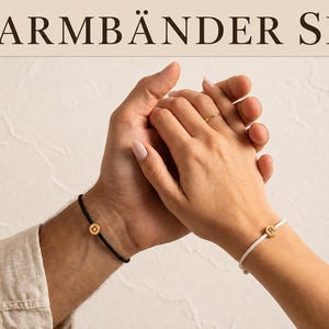 Könnte beinhalten: Ein Set aus zwei Armbändern mit dünnen Kordeln, eines in Schwarz und eines in Weiß, jeweils mit einem kleinen runden goldfarbenen Anhänger versehen. Das schwarze Armband zeigt den Buchstaben D, das weiße den Buchstaben R. Text: 2 ARMBÄNDER SET.