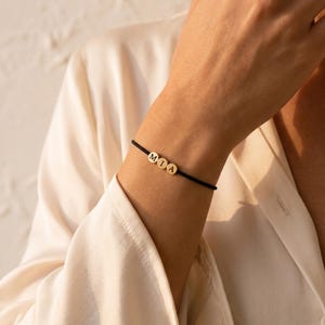 Könnte beinhalten: Ein dünnes schwarzes Kordelarmband mit drei kleinen, runden, goldfarbenen Perlen. Jede Perle ist mit einem einzelnen schwarzen Buchstaben graviert, die zusammen den Namen MIA in einer horizontalen Reihe auf dem Band bilden.